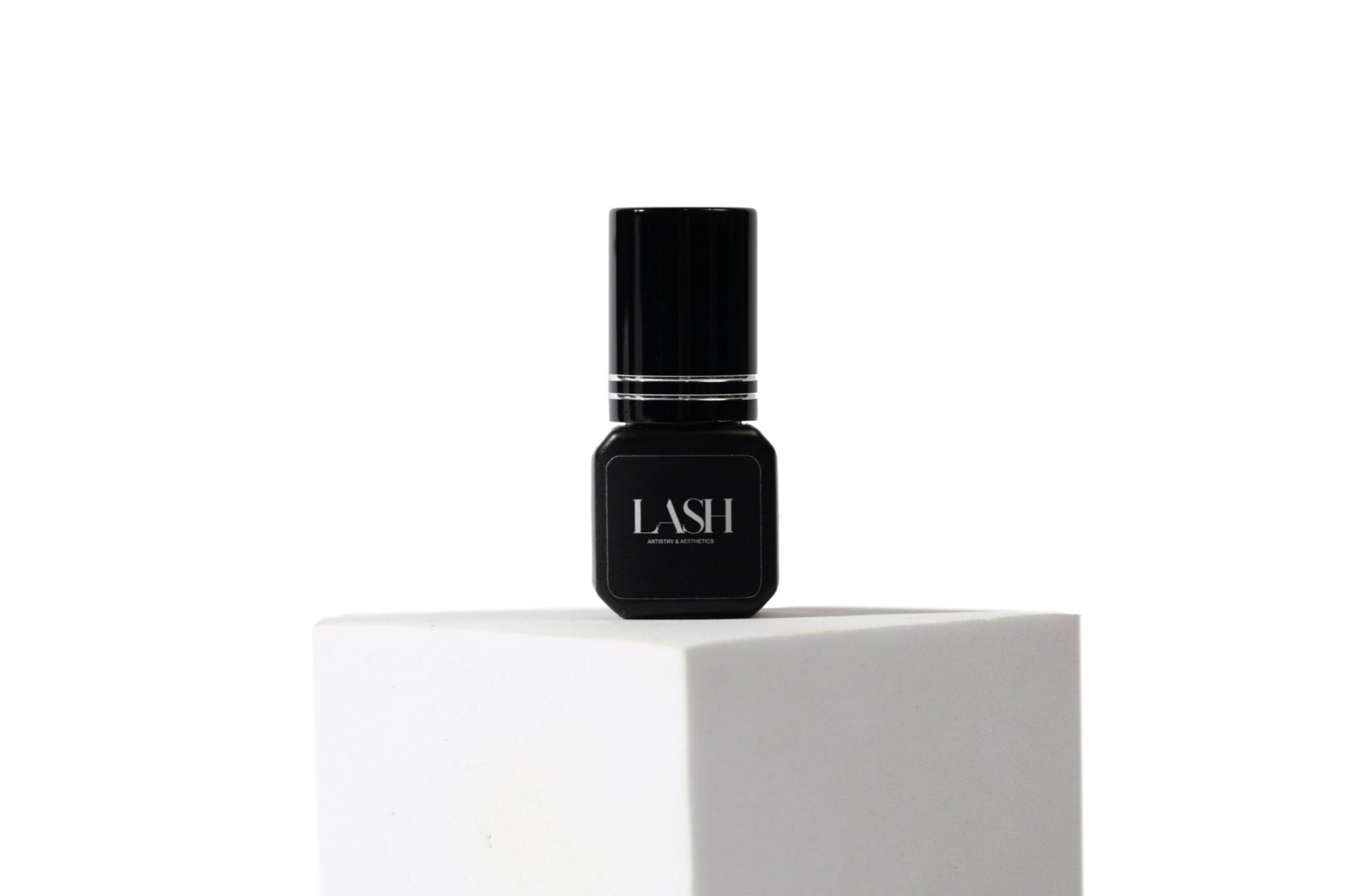 A small black bottle of lash glue on a white pedestal, with a clear label displaying the brand name 'LASH'.